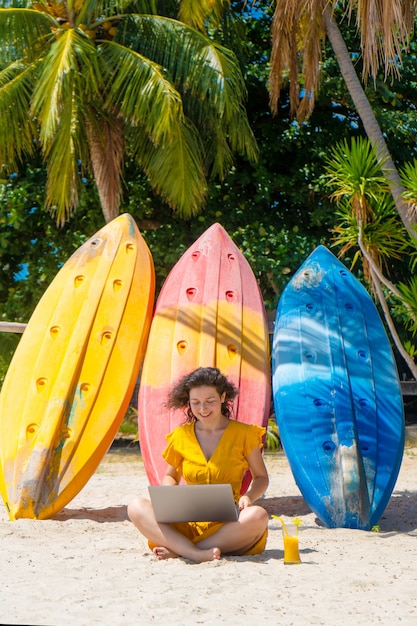 La ragazza in un vestito giallo su una spiaggia sabbiosa tropicale lavora ad un computer portatile vicino ai kajak e beve il mango fresco. Lavoro a distanza, libero professionista. Funziona in vacanza.