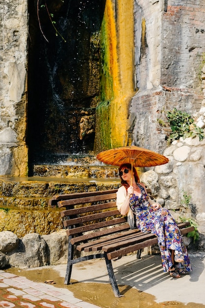 La ragazza in un vestito lungo è seduta su una panca di legno con un ombrello cinese