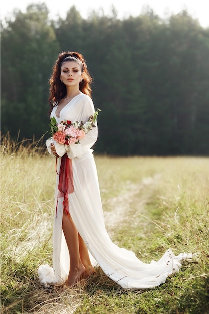 La ragazza in un vestito lungo sta in un campo con la corona sulla sua testa e un mazzo di fiori nelle sue mani, bella donna sotto i raggi del sole della sera in autunno nel villaggio. Vita rurale e moda