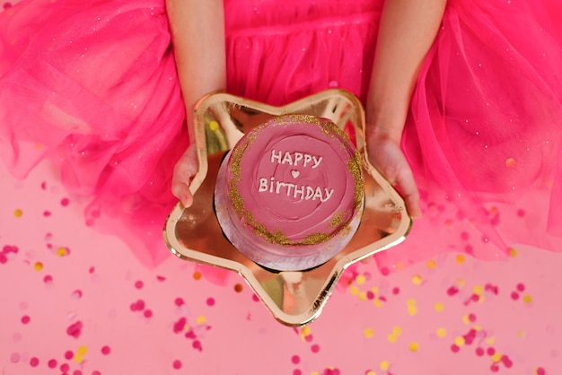 La ragazza in un vestito rosa tiene tra le mani una deliziosa torta bento dolce con la scritta buon compleanno su un piatto d'oro a forma di stella Deserto alla moda porzionato per una persona