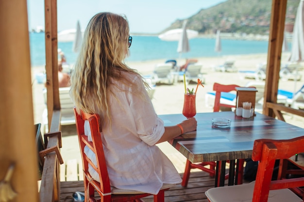La ragazza in una camicia bianca si siede con un cocktail in un ristorante vicino al mare