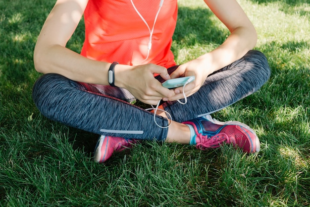 La ragazza in vestiti di sport ascolta musica sulle cuffie che si siedono sull&#39;erba nel parco dell&#39;estate