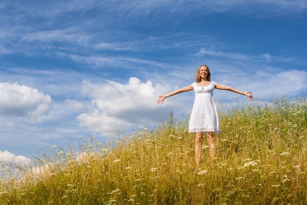 La ragazza in vestito bianco sta stando sulla collina dell'erba sul fondo del cielo blu nel giorno di estate.