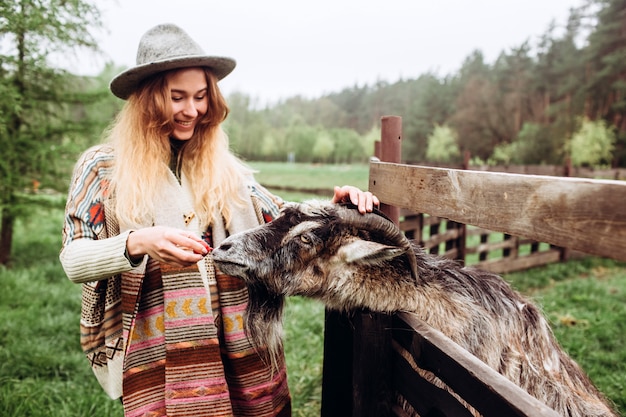 La ragazza in vestito etnico alimenta il bestiame in un'azienda agricola. Una donna in jeans un maglione di lana e un cappello grigio alimenta una capra in un vecchio ranch. Contatta il mini zoo all'aperto.