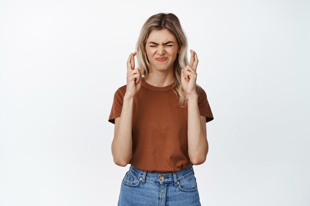 La ragazza incrocia le dita per buona fortuna, esprime un desiderio con gli occhi chiusi, pregando per smth, in piedi in maglietta e jeans su bianco.