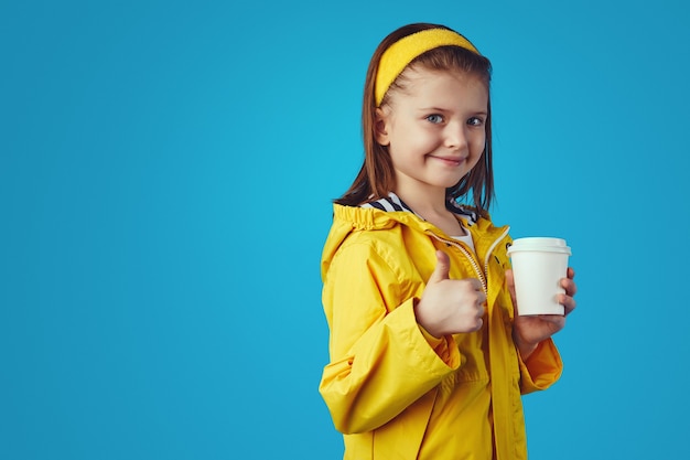 La ragazza indossa un impermeabile tiene una tazza da asporto con il tè e mostra il pollice in su