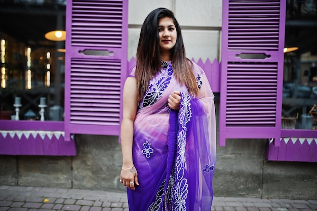 La ragazza indù indiana al tradizionale saree viola ha posato in strada contro le finestre viola