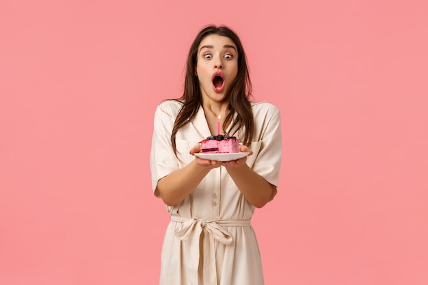 La ragazza inspira aria, respira profondamente per spegnere la candela di compleanno e far avverare il desiderio. Femmina allegra attraente in vestito che celebra, avendo festa di compleanno, tenendo la torta che sta parete rosa