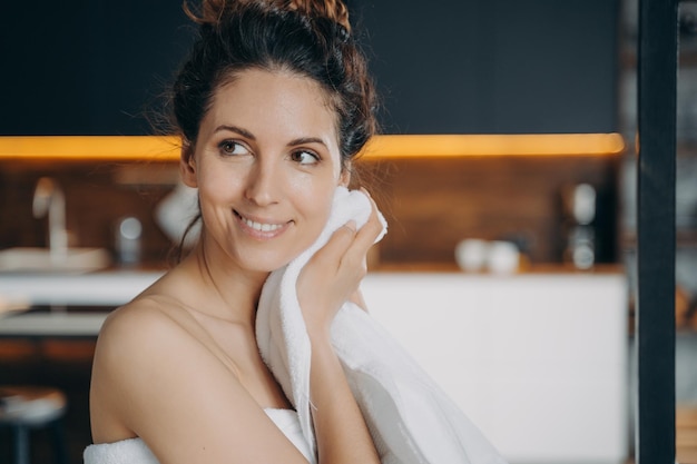 La ragazza ispanica sorridente che pulisce il viso con un asciugamano gode di una pelle fresca e luminosa dopo il trattamento per la cura della pelle a casa
