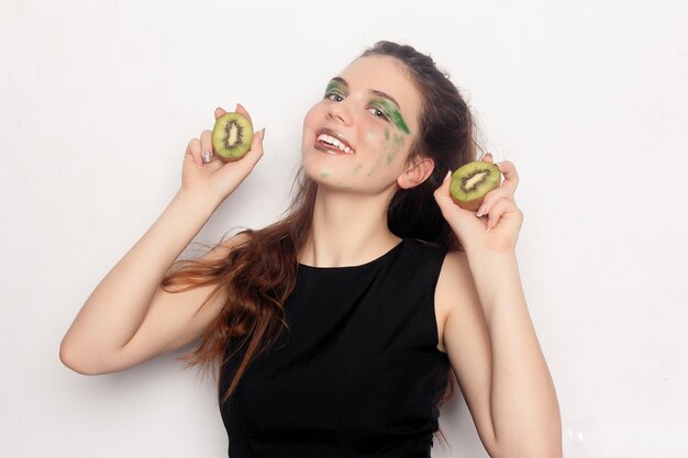 La ragazza mangia mezzo kiwi. Felice giovane bella estatica bella giovane donna che tiene due kiwi tagliati sugli occhi per uno scherzo divertente o vitamina C dinamica, al chiuso
