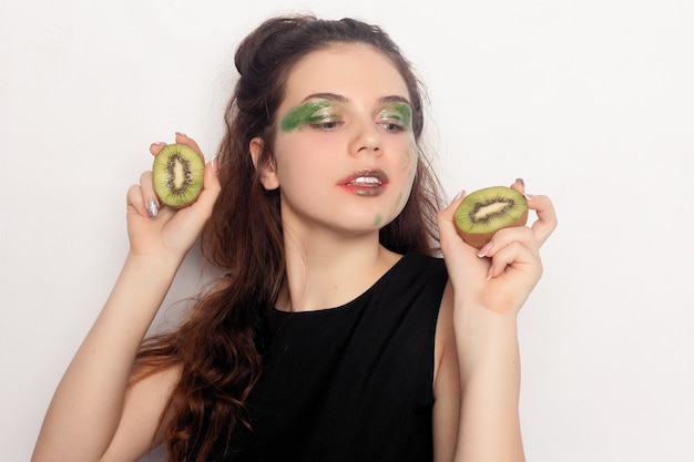 La ragazza mangia mezzo kiwi. Felice giovane bella estatica bella giovane donna che tiene due kiwi tagliati sugli occhi per uno scherzo divertente o vitamina C dinamica, al chiuso