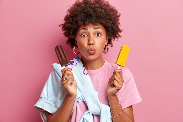 La ragazza millenaria adorabile e affettuosa dai capelli ricci si gode la dolce estate tiene le labbra piegate vuole baciare qualcuno tiene due gelati di cioccolato e sapore di mango indossa abiti casual pose al coperto