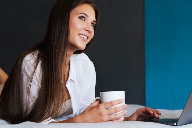 La ragazza millenaria sta riposando sul letto in camicia bianca, tenendo la tazza di caffè in mano.