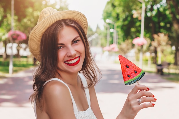 La ragazza nel buon umore cammina nel parco e sorride. Dolce ragazza felice in cappello di paglia passeggiate nel parco con una lecca-lecca a forma di anguria