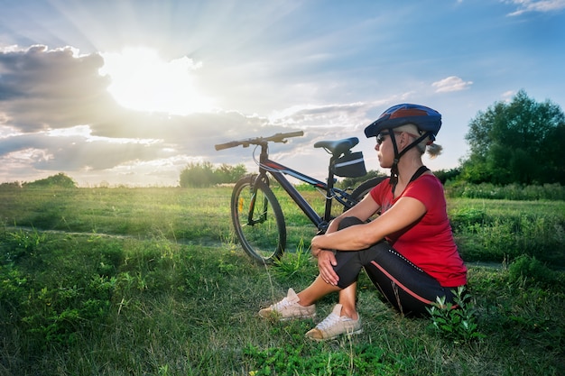 La ragazza nel casco da bicicletta si siede vicino alla bicicletta e guarda il tramonto