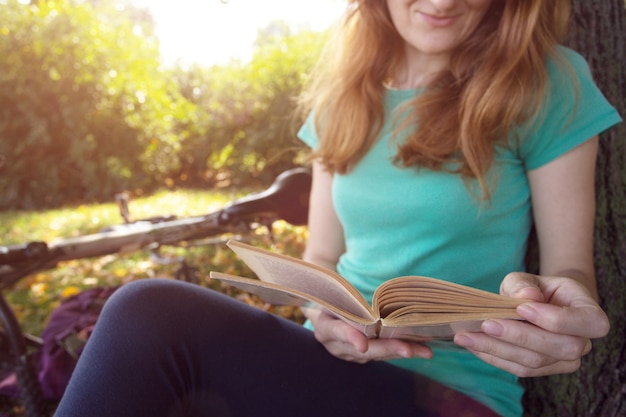 La ragazza nel parco si siede sotto un albero e legge un libro. accanto c'è una bicicletta e uno zaino