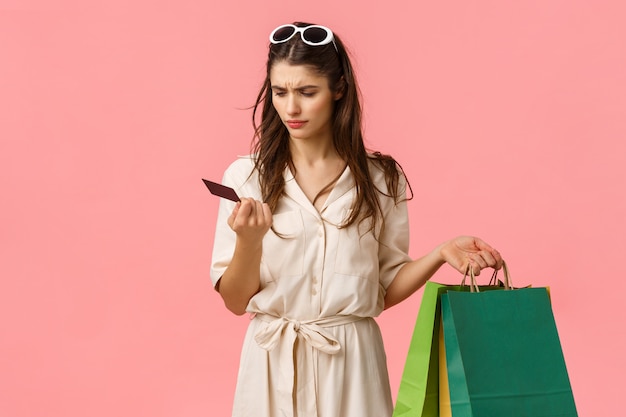 La ragazza non capisce dove vanno tutti i soldi. Shopaholic femminile perplesso e confuso che tiene i sacchetti della spesa e che fissa seriamente alla carta di credito, stando parete rosa all'oscuro