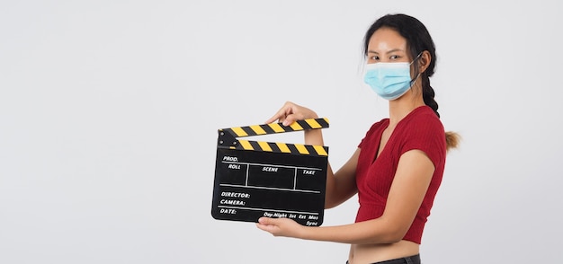 La ragazza o la donna asiatica indossano la maschera per il viso e la mano che tiene il ciak o l'ardesia del film nella produzione di video, film, industria cinematografica su sfondo bianco.