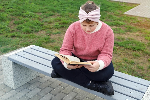La ragazza paffuta si siede sulla panchina e legge premurosa il libro nel parco