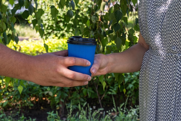 La ragazza passa il bicchiere di caffè blu al ragazzo di mano in mano foglie sfocate di un albero nel