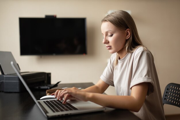 La ragazza pensierosa sveglia sta facendo il lavoro dal testo digitante domestico su un computer con un taccuino e un telefono sulla tavola e sulla pandemia.