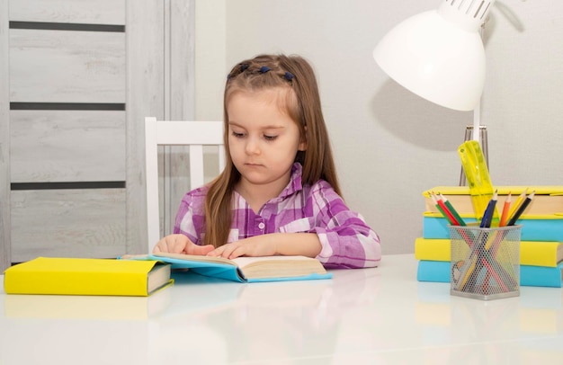 La ragazza piccola e affascinante è seduta al tavolo con i libri, preparandosi per la scuola. Messa a fuoco selettiva.