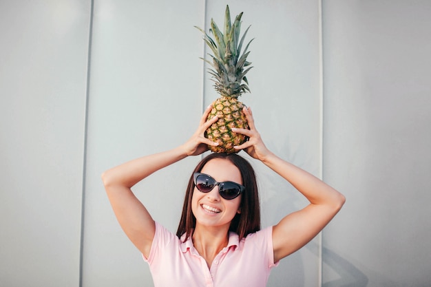 La ragazza positiva in occhiali da sole sta e tiene l'ananas sulla sua testa. Lo mantiene in equilibrio con le mani.