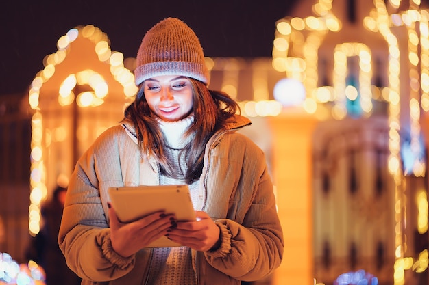 La ragazza positiva usa la sua tavoletta digitale mentre sta in piedi sullo sfondo delle vacanze di Natale all'aperto