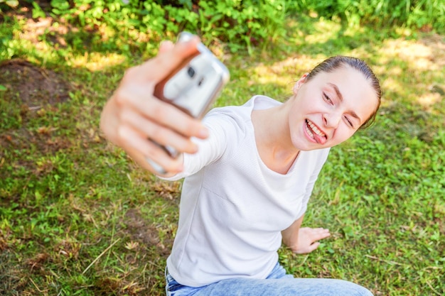 La ragazza prende selfie dalle mani con il telefono che mostra la lingua e la faccia buffa che si siede sullo sfondo del parco o del giardino. Ritratto di giovane donna attraente che fa foto selfie su smartphone nel giorno d'estate