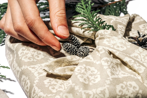 La ragazza prepara la scatola regalo fatta a mano beige di Natale per il Natale con il thuja ed il cono del tessuto. Confezione regalo con zero rifiuti in stile furoshiki tradizionale giapponese. Concetto ecologico arredamento ecologico natale. Macro.