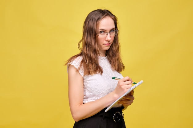 La ragazza rigorosa dell'insegnante con i vetri su un fondo giallo prende nota con una penna in un taccuino.