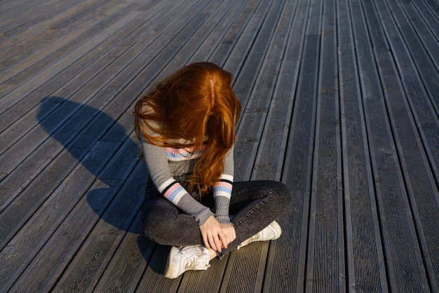 La ragazza rossa triste si siede su un pavimento di legno nel parco in una giornata di sole