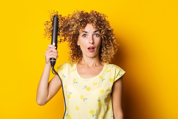 La ragazza scioccata con i capelli ricci si raddrizza i capelli con una piastra per capelli su uno sfondo giallo