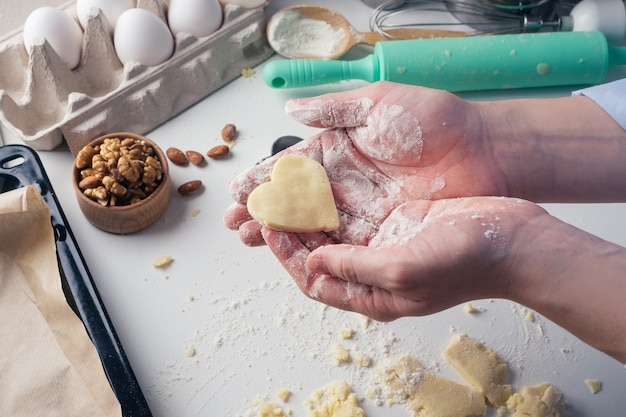 La ragazza scolpisce i biscotti a forma di cuore in cucina, a casa, primo piano. Dolci fatti a mano con amore per San Valentino, festa della mamma o festa del papà. Sfondo culinario. Cibo per le vacanze.