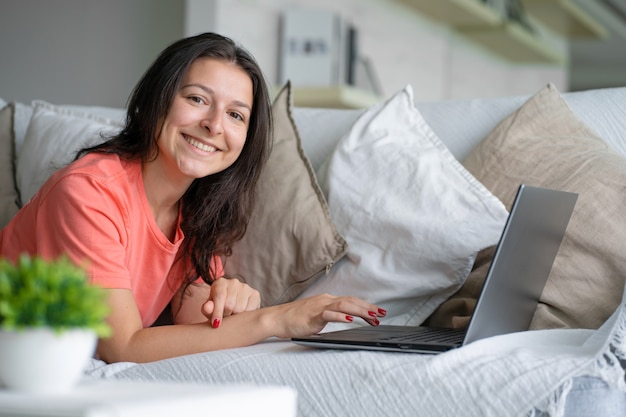 La ragazza sdraiata sul divano si rallegra guardando il laptop. Sorrisi, buon umore, emozione di gioia.