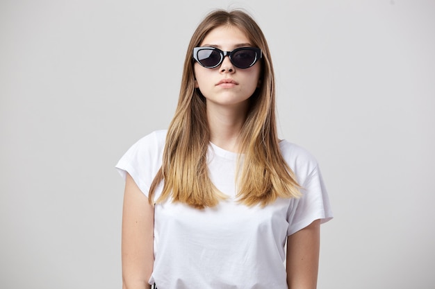 La ragazza seria vestita con una maglietta bianca e occhiali da sole è su uno sfondo bianco in studio.