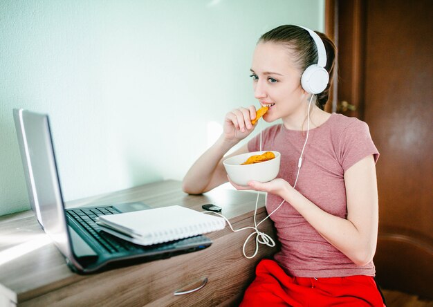 La ragazza si siede ad un laptop nelle cuffie, ride e mangia i chip