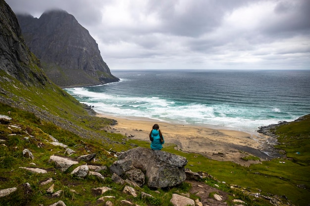 la ragazza si siede su una roccia godendosi la vista dalla cima della spiaggia di kvalvika sulle isole lofoten, norvegia