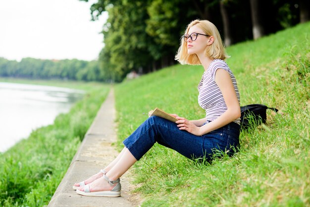 La ragazza si siede sull'erba verde vicino al fiume Stagione delle vacanze Vacanze estive Ragazza che si rilassa in riva al fiume