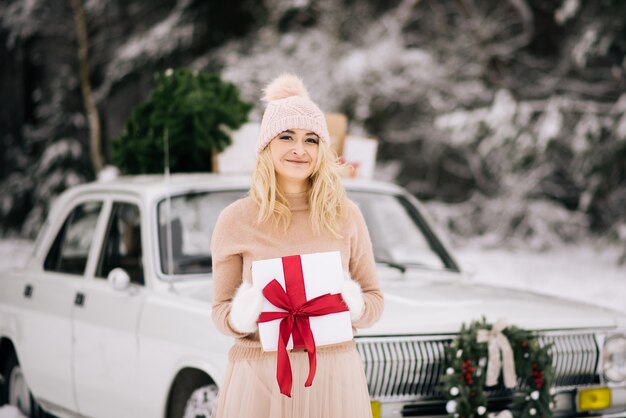 La ragazza si sta preparando per il Natale, si leva in piedi con un regalo in mano sullo sfondo di un'auto retrò, il cui tetto è un albero di Natale, regali e una corona nella foresta innevata invernale.