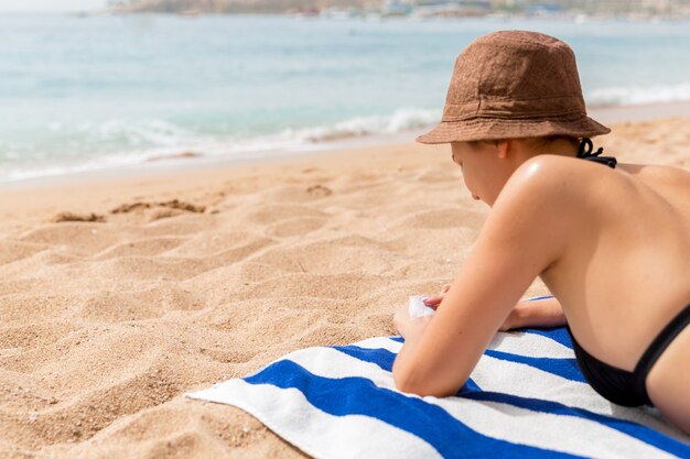 La ragazza si sta rilassando sull'asciugamano sulla sabbia in spiaggia e sta applicando una crema solare sulla mano.