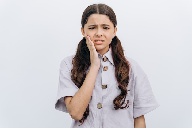 La ragazza si tocca la guancia con la mano, sussulta per il mal di denti. La bambina sveglia sussultò per il dolore su uno sfondo bianco