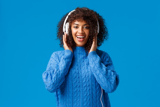 La ragazza sorridente afroamericana carismatica felice ha ottenuto le nuove cuffie del regalo di Natale, ascolta musica e gode di battiti fantastici, tocca gli auricolari e guarda la fotocamera, blu