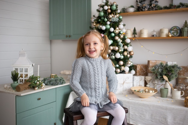 La ragazza sorridente del bambino sta sedendosi nella cucina vicino all'albero di Natale decorato all'interno.