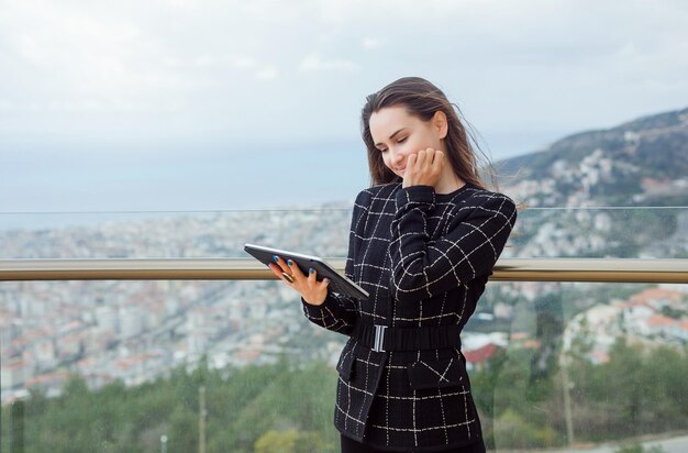 La ragazza sorridente del blogger sta guardando il computer Planshet in mano tenendo la mano sulla guancia sullo sfondo della vista della città