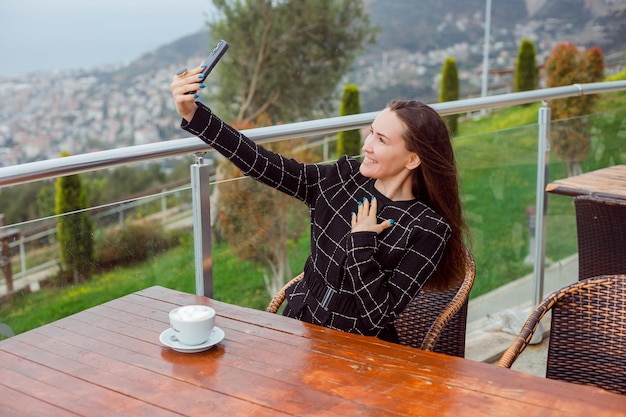 La ragazza sorridente del blogger sta prendendo selfie con il suo smartphone tenendo la mano sul petto sedendosi sullo sfondo della vista della città