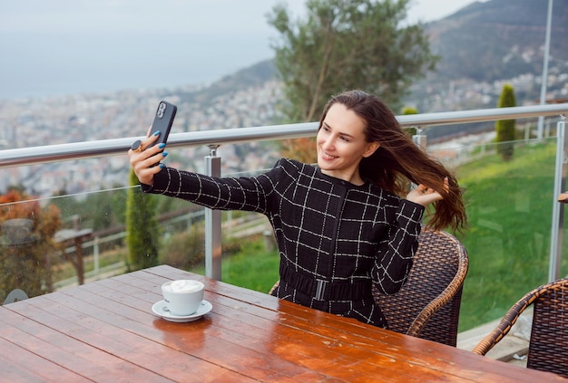 La ragazza sorridente del blogger sta prendendo selfie con lo smartphone tenendosi i capelli seduto su uno sfondo con vista sulla città