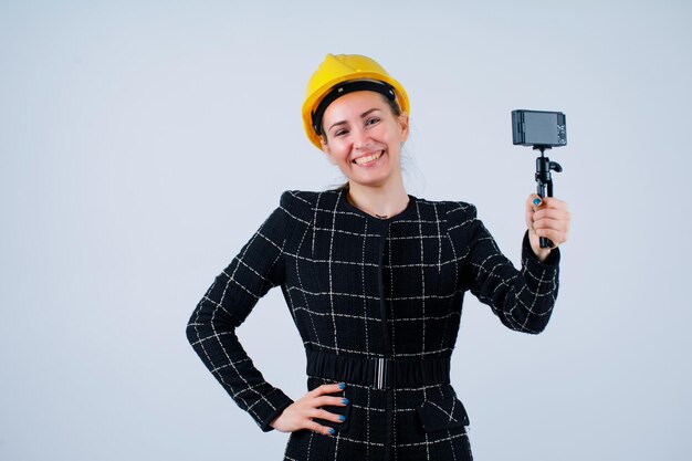 La ragazza sorridente dell'ingegnere sta tenendo la sua mini macchina fotografica e sta mettendo l'altra mano sulla vita su fondo bianco