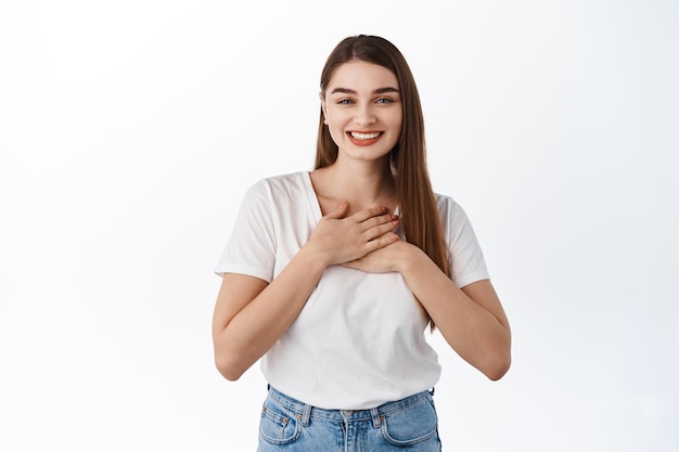 La ragazza sorridente e grata apprezza il bel gesto, tenendosi per mano sul cuore grato, dire grazie, stando in piedi sincera e commossa su sfondo bianco