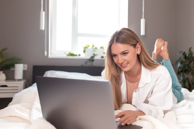 La ragazza sorridente giace sul letto in camera da letto e utilizza il laptop per il lavoro a distanza